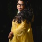 A woman in glasses and yellow traditional dress, posing gracefully.