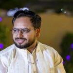 A man in traditional attire smiles with colorful bokeh lights in the background, creating a festive mood.
